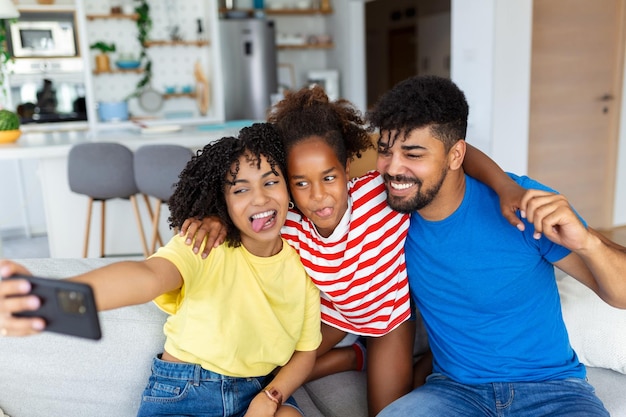 Familia joven sonriente con su hija sentada en el sofá en la cocina hacen una foto de autorretrato en la celda juntos padres felices con un niño pequeño se divierten toman selfie en el teléfono inteligente en casa