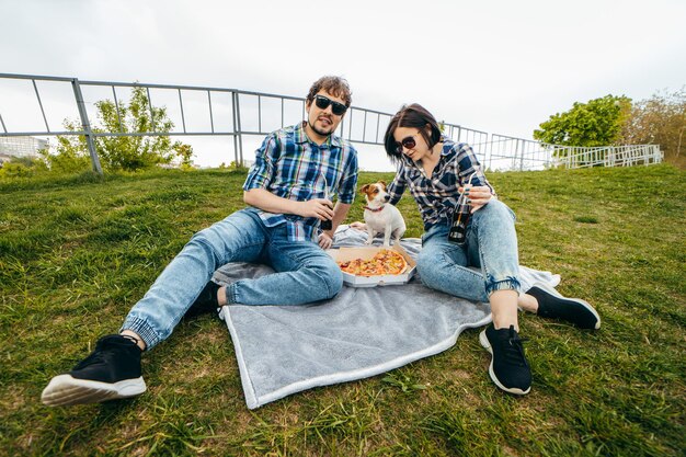 Foto familia joven con su perro sentado en el césped y comiendo pizza al aire libre