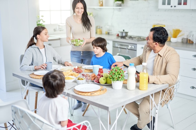 Família latina feliz jantando juntos em casa. Mulher alegre, sorrindo enquanto servia salada para o marido e os filhos, em pé na cozinha. Foco seletivo