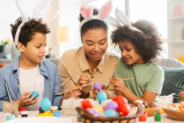 Familia latina sonriente madre e hijos con orejas de conejo pintando huevos de Pascua en casa