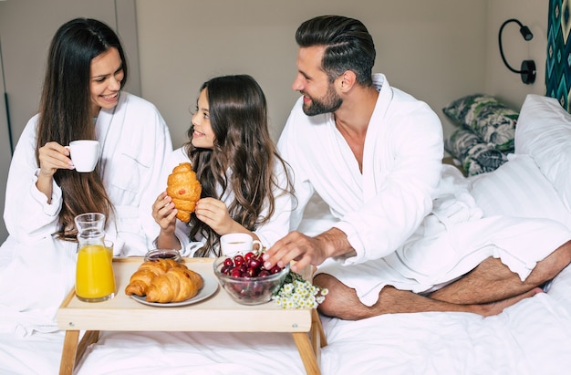 Família linda e feliz em roupões de banho tomando café da manhã em um quarto de hotel de luxo