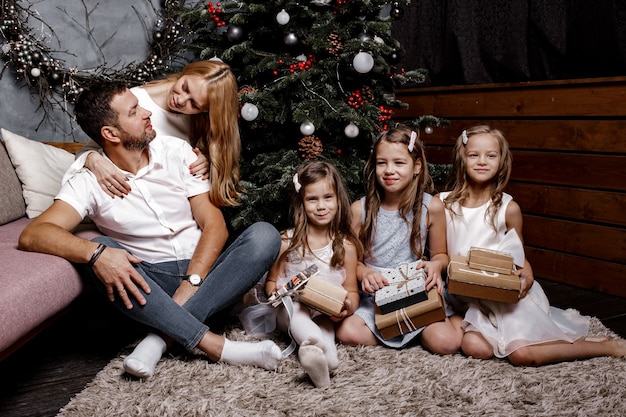 Familia linda feliz con tres niños intercambiando regalos bajo el árbol de Navidad en la alfombra.