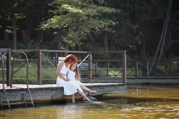 Familia, madre e hija están sentadas junto al río. Niña con su mamá. Chicas con vestidos blancos. Tiempo en familia juntos.