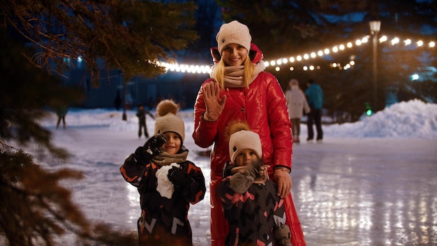 Una familia de madre joven y dos niños parados en la pista de hielo decorada y saludando con las manos