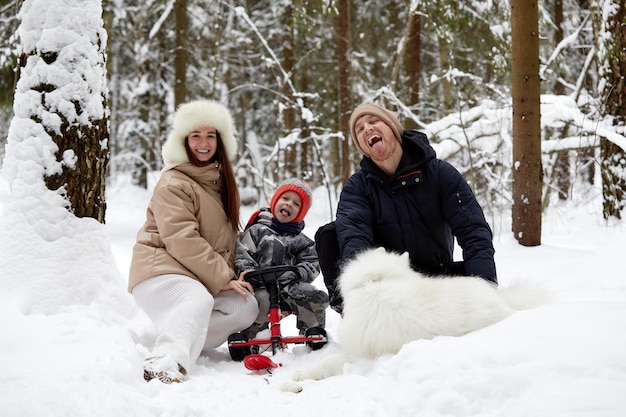 Familia de madre, padre e hijo divirtiéndose en madera de invierno nevada con perro mascota alegre