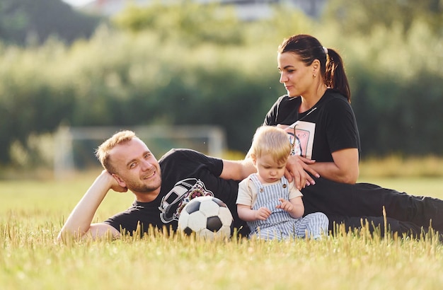 Familia de madre padre y niño pequeño está en el campo verde con la pelota de fútbol