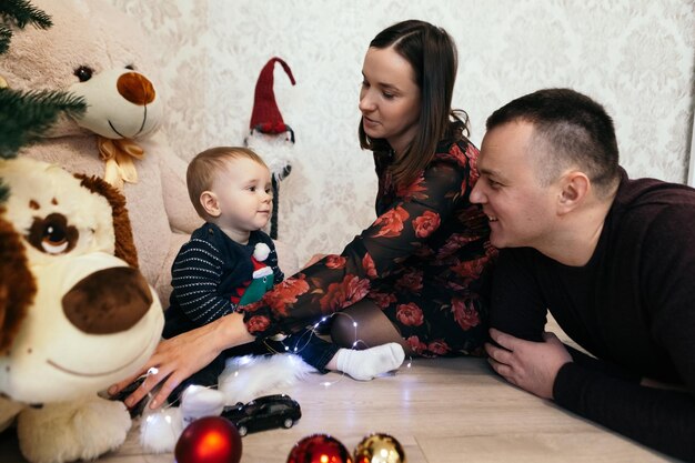 Foto familia madre padre niño pequeño niño lindo niño juguetón niño pequeño sentado cerca del árbol de navidad