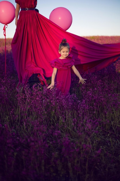 Família mãe e filha em vestidos rosa estão de pé em um campo com flores e grandes balões ao pôr do sol no verão contra o céu azul