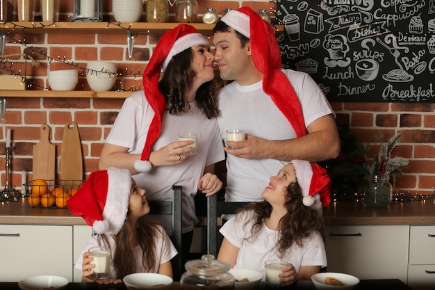 Foto família, mãe, pai e 2 filhas na cozinha usando chapéus de papai noel com leite nas mãos