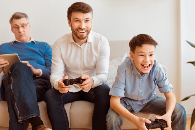 Familia masculina fin de semana padre e hijo jugando a videojuegos sentados junto al abuelo en el sofá en casa