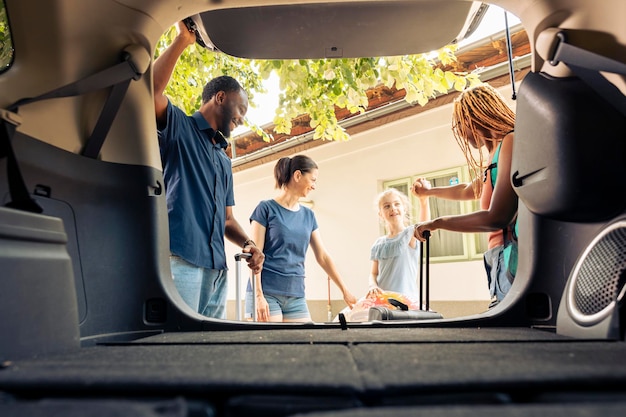 Familia mixta y amigos que viajan de vacaciones con vehículo, yendo de vacaciones de verano. Niña con madre y pareja joven saliendo con automóvil en viaje por carretera, aventura.