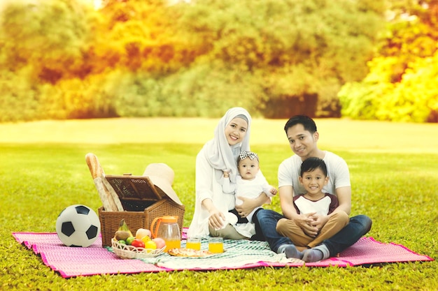 Família muçulmana feliz olhando para a câmera enquanto fazia piquenique no parque com fundo de árvores de outono