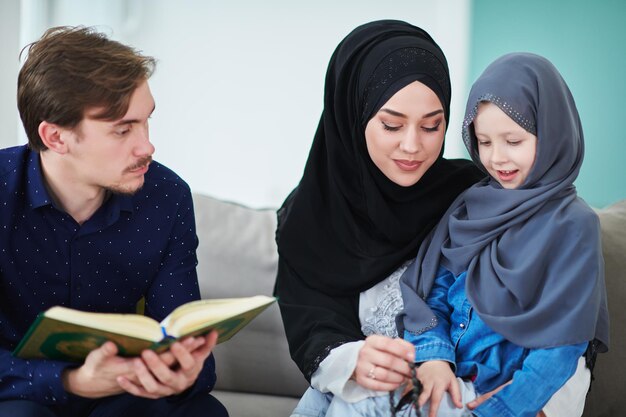 Família muçulmana jovem lendo o Alcorão durante o Ramadã. Pais e filha adorando a Deus, em roupas islâmicas em casa moderna