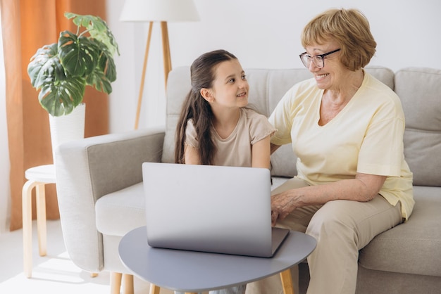 Familia multigeneracional abuela y nieta se sientan en el sofá con computadora inalámbrica se miran con ternura y amor disfrutan del ocio en Internet y el tiempo juntos ven películas en línea