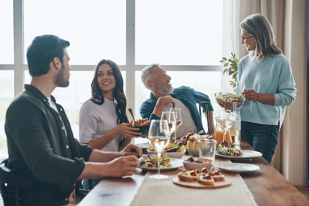 Familia multigeneracional moderna disfrutando de la cena y sonriendo mientras está sentado en el apartamento moderno