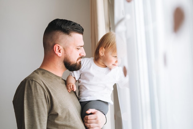 Familia multinacional Joven padre con una niña en el alféizar de la ventana mirando la ventana en casa