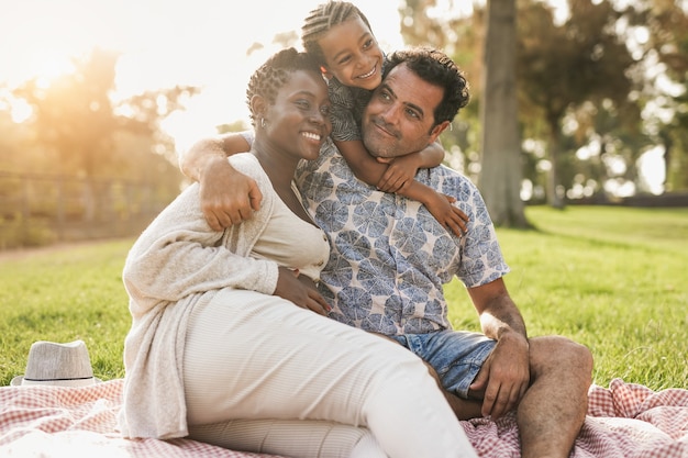 Família multirracial feliz se abraçando no parque da cidade - Mulher grávida africana com marido caucasiano e criança mestiça fazem piquenique ao ar livre