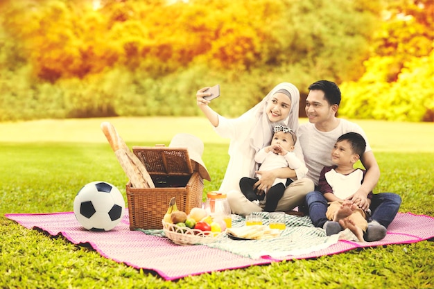 Familia musulmana usando un teléfono celular para tomar una foto selfie juntos mientras hacen un picnic en el parque con fondo de árboles otoñales