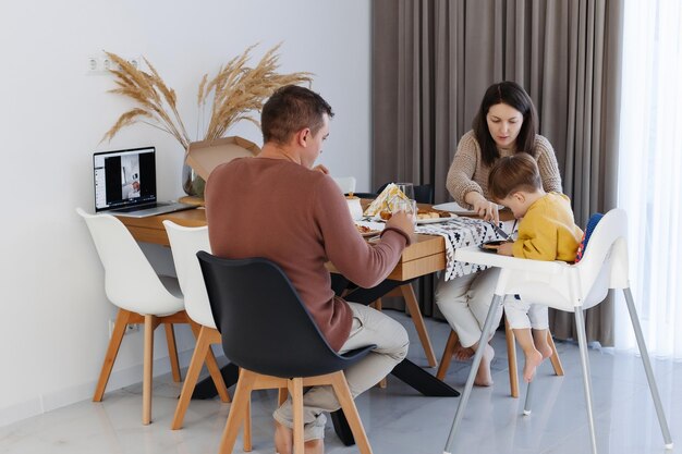 Família na cozinha comendo pizza perto do computador em que a transmissão de vídeo de uma criança