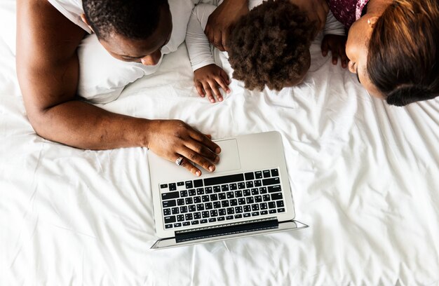 Familia negra acostada en la cama usando una computadora portátil juntos en el dormitorio