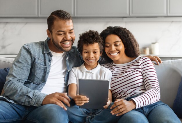 Foto família negra com filho pré-adolescente sorrindo alegremente enquanto usam um tablet digital juntos