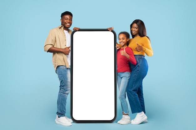 Familia negra de pie cerca de un teléfono inteligente gigante con pantalla en blanco que muestra los pulgares hacia arriba y anuncia una aplicación o sitio web genial