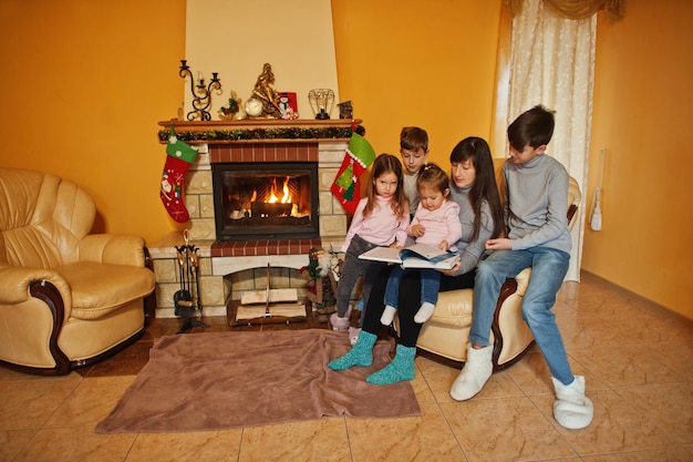 Familia numerosa joven feliz junto a una chimenea en la cálida sala de estar en día de invierno. Madre con cuatro hijos en casa lee el libro.