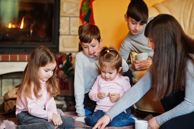 Familia numerosa joven feliz junto a una chimenea en la cálida sala de estar en día de invierno. Madre con cuatro hijos en casa lee el libro.