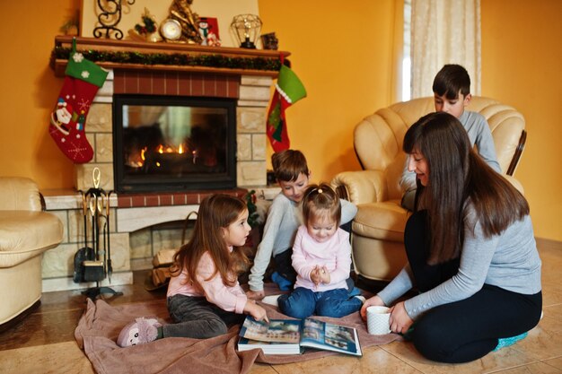 Familia numerosa joven feliz junto a una chimenea en la cálida sala de estar en día de invierno. Madre con cuatro hijos en casa lee el libro.