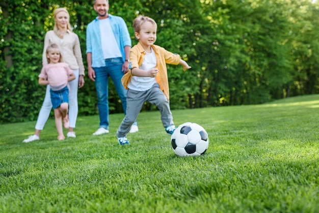 família olhando garotinho brincando com bola de futebol no parque