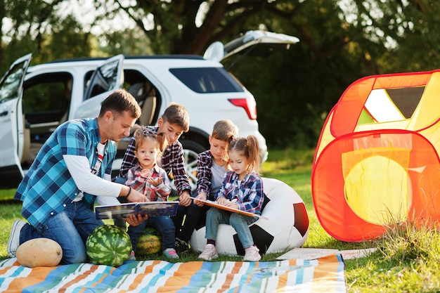 Familia pasando tiempo juntos Padre leyendo un libro al aire libre con niños contra su auto todoterreno