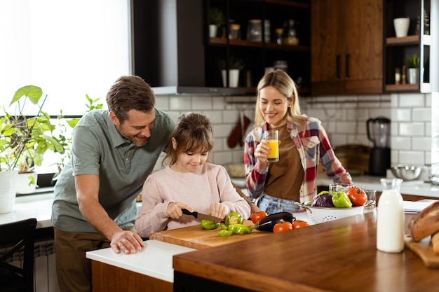 La familia prepara una comida juntos con el padre ayudando a la hija a cortar verduras y la madre disfrutando de un jugo