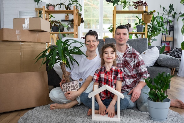 Família que se mudou com um filho e uma planta doméstica está desfrutando de uma nova casa sentada no sofá e segurando o telhado perto de caixas com coisas Compra de seguro hipotecário e mudança para sua casa