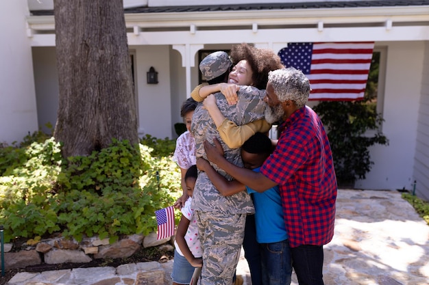 Familia de raza mixta de varias generaciones disfrutando de su tiempo en un jardín, dando la bienvenida a un hombre afroamericano con uniforme militar, regresando a casa, abrazando a su familia, en un día soleado