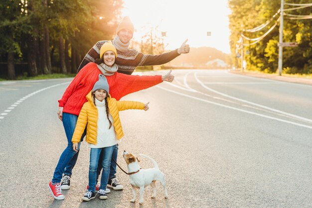 Una familia relajada y despreocupada camina por la carretera en el campo con un perro que levanta los pulgares con alegría, usa ropa abrigada y calzado deportivo, ya que tiene un largo camino Gestos familiares jóvenes que tienen tiempo libre