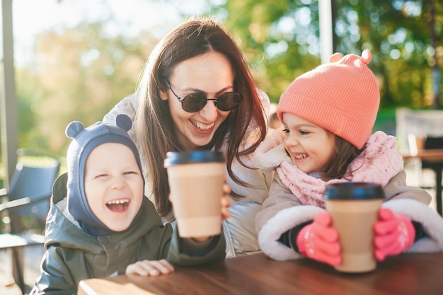 Família rindo, uma mãe com dois filhos pequenos se divertindo juntos em um café ensolarado ao ar livre em uma estação fria