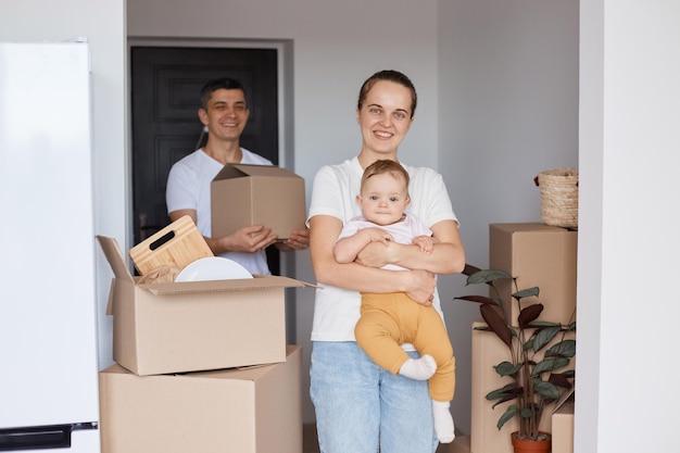 Familia satisfecha, madre, padre e hija pequeña posando en un nuevo apartamento después de mudarse, expresando felicidad rodeada de muchas cosas, reubicación, renovación, mudanzas y servicio de entrega.