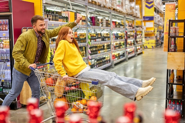 Família se diverte no corredor do supermercado, mulher senta no carrinho e gosta de fazer compras com o marido. vista lateral