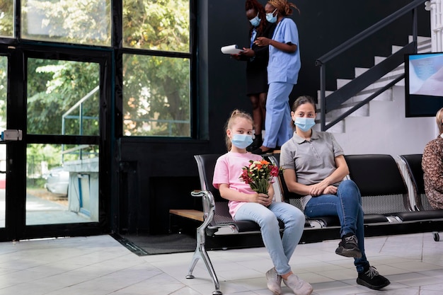 Familia sentada en una silla en la sala de espera del hospital durante la consulta de la visita de control, usando mascarilla médica para prevenir la infección con covid19. Niña con ramo de flores. concepto de medicina