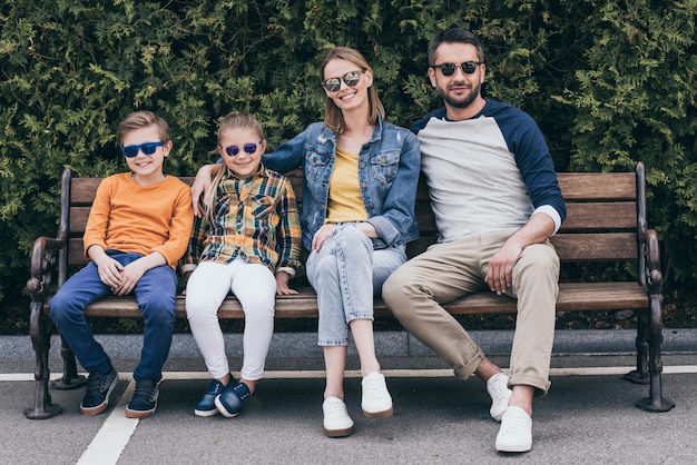 familia sonriente con gafas de sol sentados juntos en un banco en el parque