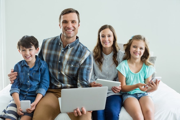 Familia sonriente con ordenador portátil, tableta digital y teléfono móvil en el dormitorio