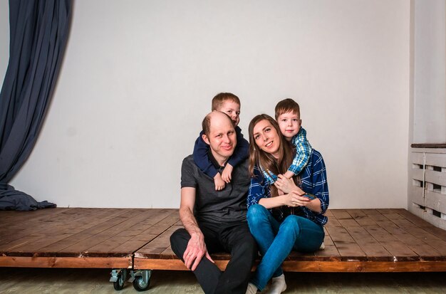 Familia sonriente sosteniendo la forma de la casa mientras está sentado en el piso de madera en casa. Sesión de fotos de una familia con 2 hijos, fondo blanco. Relaciones familiares cálidas. Padres jovenes