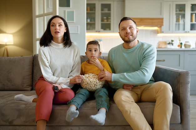Familia sorprendida viendo una película comiendo palomitas de maíz sentada en un sofá en la sala de estar