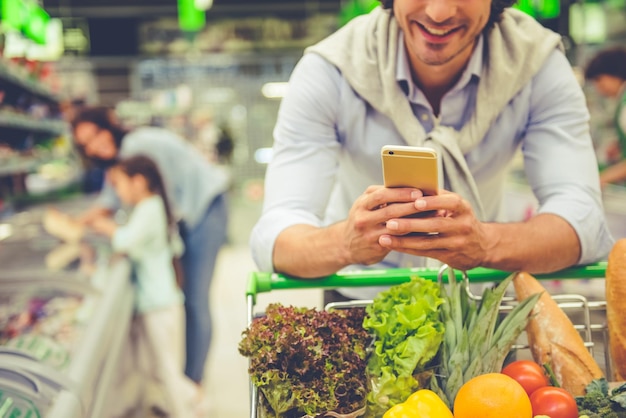 Familia en el supermercado Imagen recortada de papá apoyado en un carrito de compras usando un teléfono móvil y sonriendo en el fondo, su esposa y su hija están eligiendo comida