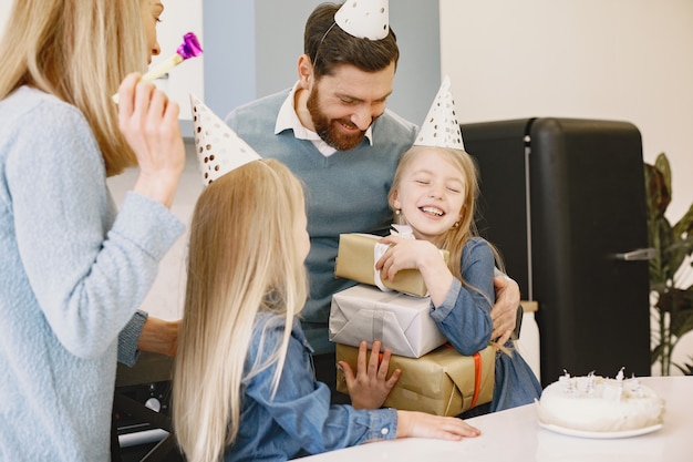 La familia y sus dos hijas celebran su cumpleaños en la cocina. La gente usa un sombrero de fiesta. Chica guarda cajas con regalos.