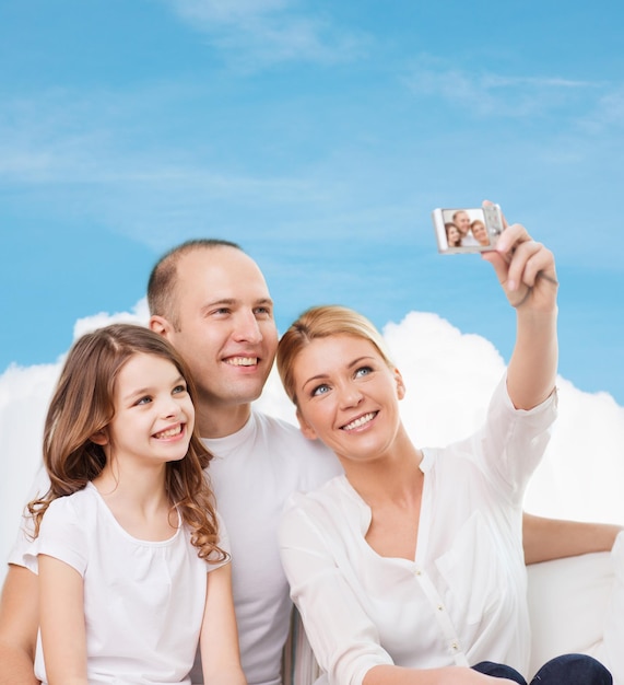 familia, tecnología y gente - madre sonriente, padre y niña haciendo selfie con cámara sobre cielo azul y fondo de nube blanca