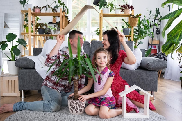 Família tradicional com criança está aproveitando a nova casa sentada no sofá e segurando o telhado Seguro hipotecário e proteção comprando e mudando para sua própria casa verde com vaso de plantas