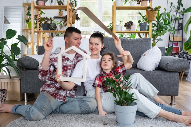 La familia tradicional con niños disfruta de un nuevo hogar sentados en el sofá y sosteniendo el techo. Seguro hipotecario y protección comprando y mudándose a su propia casa, casa verde con planta en maceta.