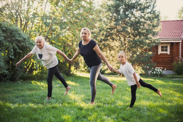 Familia de tres generaciones de pie sobre una pierna balanceándose y tomándose de la mano haciendo deportes juntos en el patio trasero en un prado lleno de hierba verde y árboles Pasando tiempo juntos