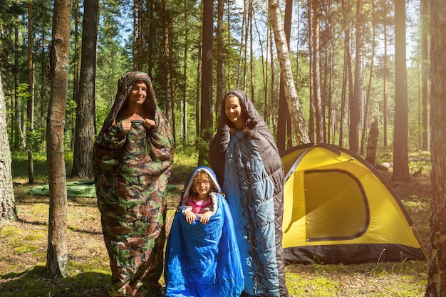Familia de turistas de padre, madre e hija pequeña posan en sacos de dormir cerca de una tienda de campaña. Recreación familiar al aire libre, turismo interno, camping, equipo de senderismo. Pupados como orugas-humor
